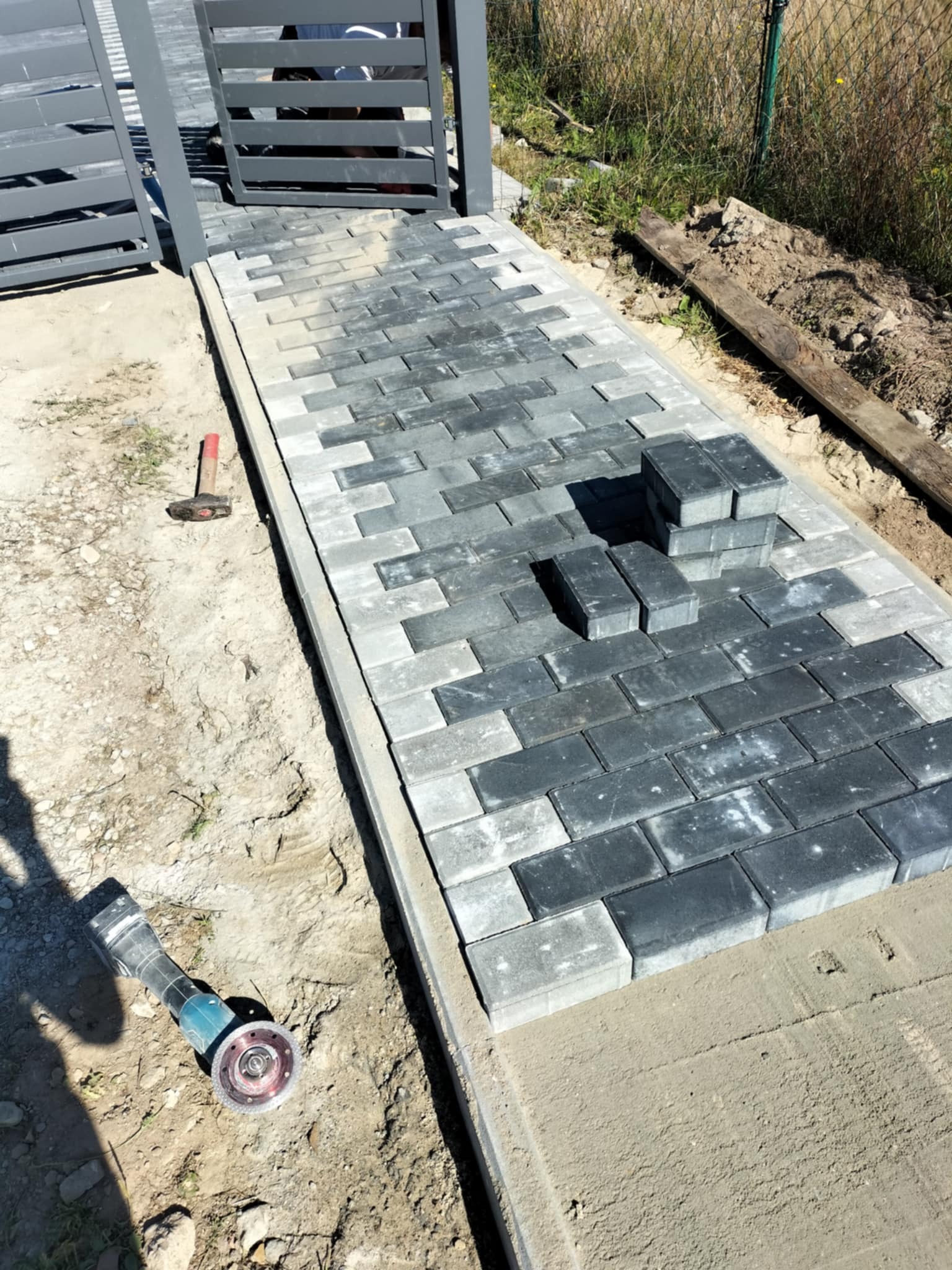 Układanie szarej kostki brukowej na ścieżce, widoczny młotek, szlifierka kątowa i fragment bramy w tle. Częściowo ułożona kostka, etap prac.