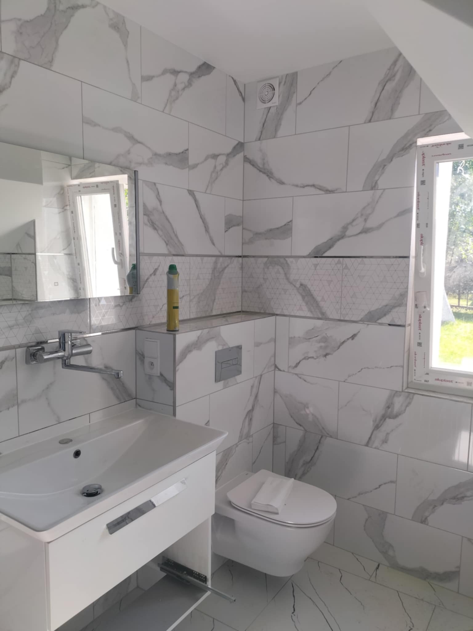 Wykończona łazienka z białymi płytkami imitującymi marmur. Umywalka z szafką, toaleta i okno z widokiem na zieleń za oknem. Na ścianie ozdobny pas z geometrycznym wzorem.