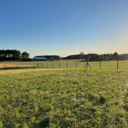 SiataNaLata.pl - Rozległy widok na łąkę z niskim ogrodzeniem z siatki na słupkach, w tle zabudowania gospodarcze i las, oświetlone wschodzącym słońcem.