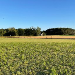 SiataNaLata.pl - Rozległy widok na polną przestrzeń z niskim, metalowym ogrodzeniem, za którym widać las i dom w oddali, pod błękitnym niebem.