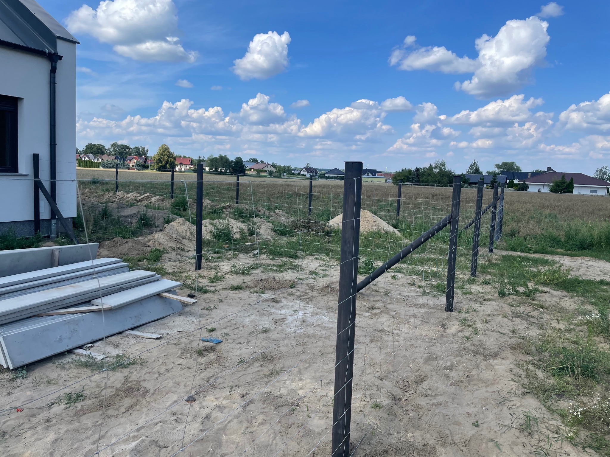 Montaż ogrodzenia z siatki i podmurówki prefabrykowanej na terenie budowy domu jednorodzinnego w słoneczny dzień. Widoczne słupki i elementy podmurówki.