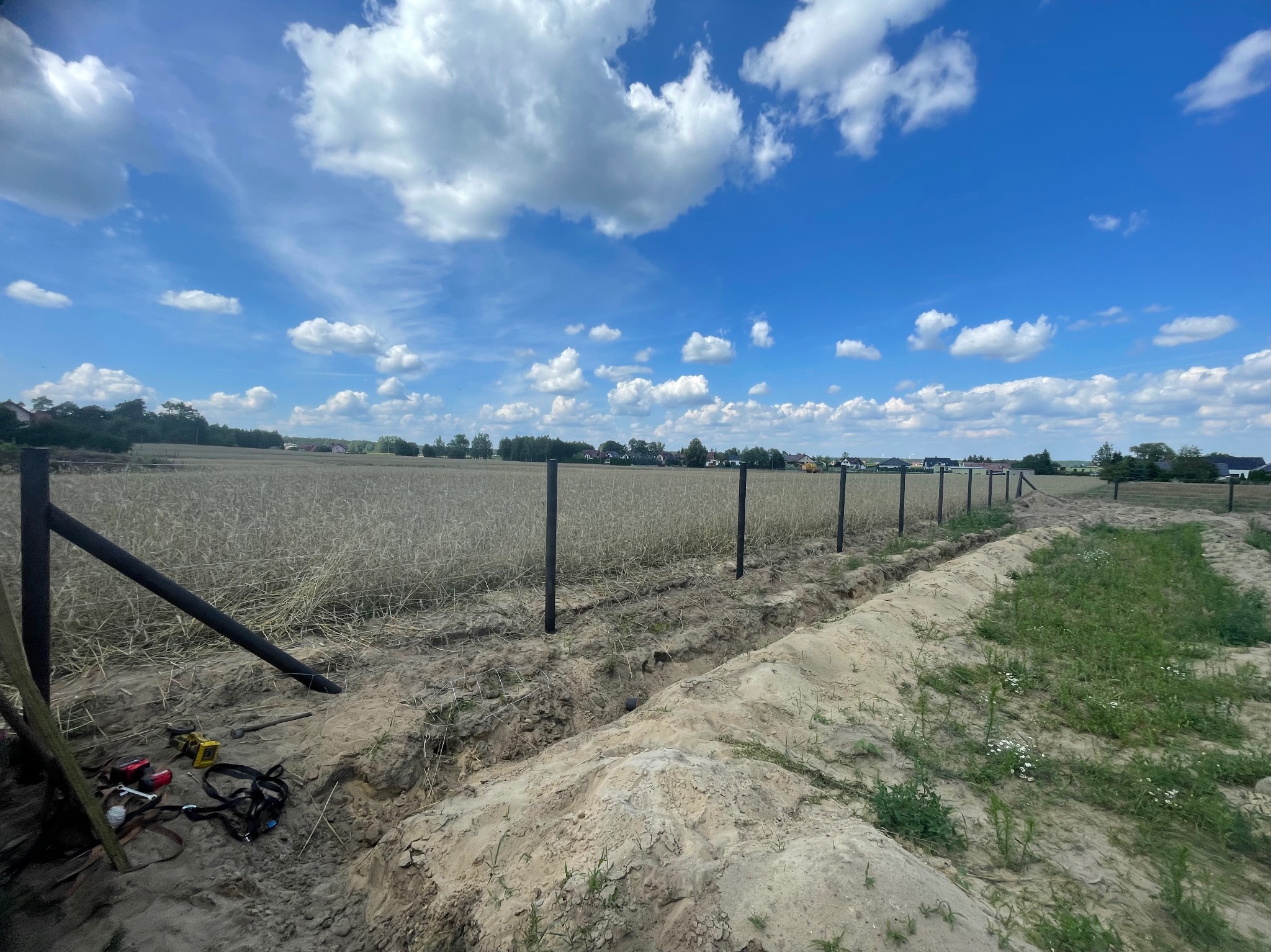 Etap instalacji ogrodzenia: czarne słupki wkopane w ziemię, wykopany rów, narzędzia obok. Pola w tle pod błękitnym niebem z chmurami.