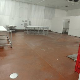 AGNIESZKA KOCIURA-SARBAK USŁUGI KOMPLEKSOWEGO SPRZĄTANIA - Wnętrze hali produkcyjnej z metalowymi stołami i brązową podłogą po myciu. Widoczne ślady wilgoci, białe ściany z blachy trapezowej i odpływ w podłodze.