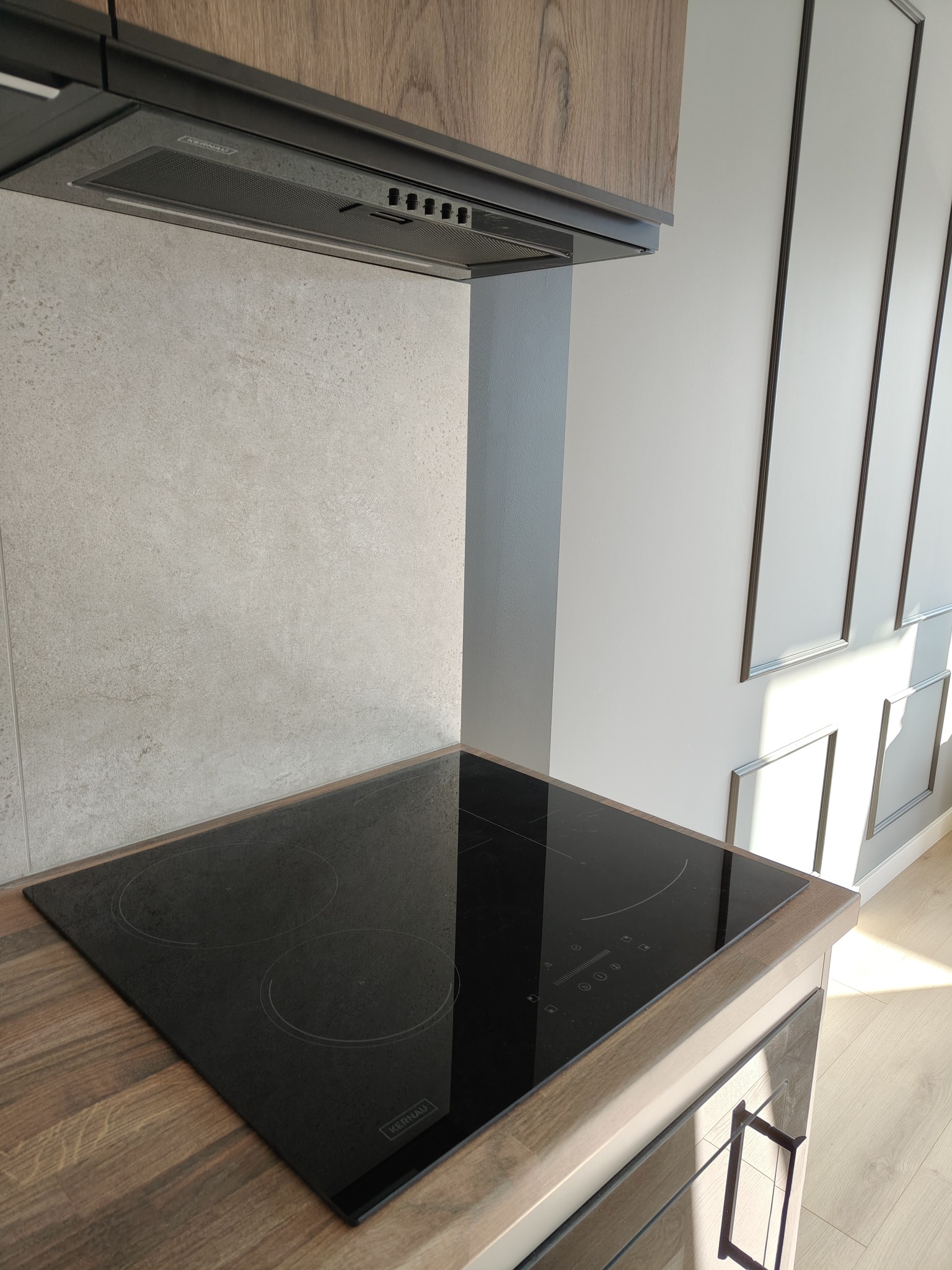 Nowoczesna płyta indukcyjna Kernau wkomponowana w drewniany blat kuchenny, z czarnym okapem i szarymi płytkami na ścianie w tle. Minimalistyczny design.