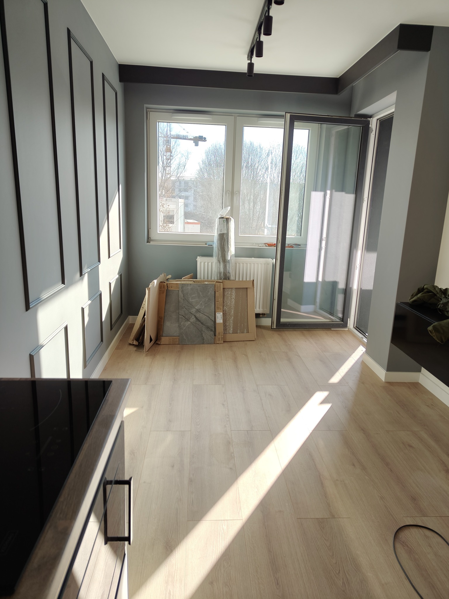 Jasne, puste wnętrze po remoncie z panelami na podłodze, szarymi ścianami i otwartym oknem balkonowym w słoneczny dzień. Widoczne kartony i płytki pod oknem.