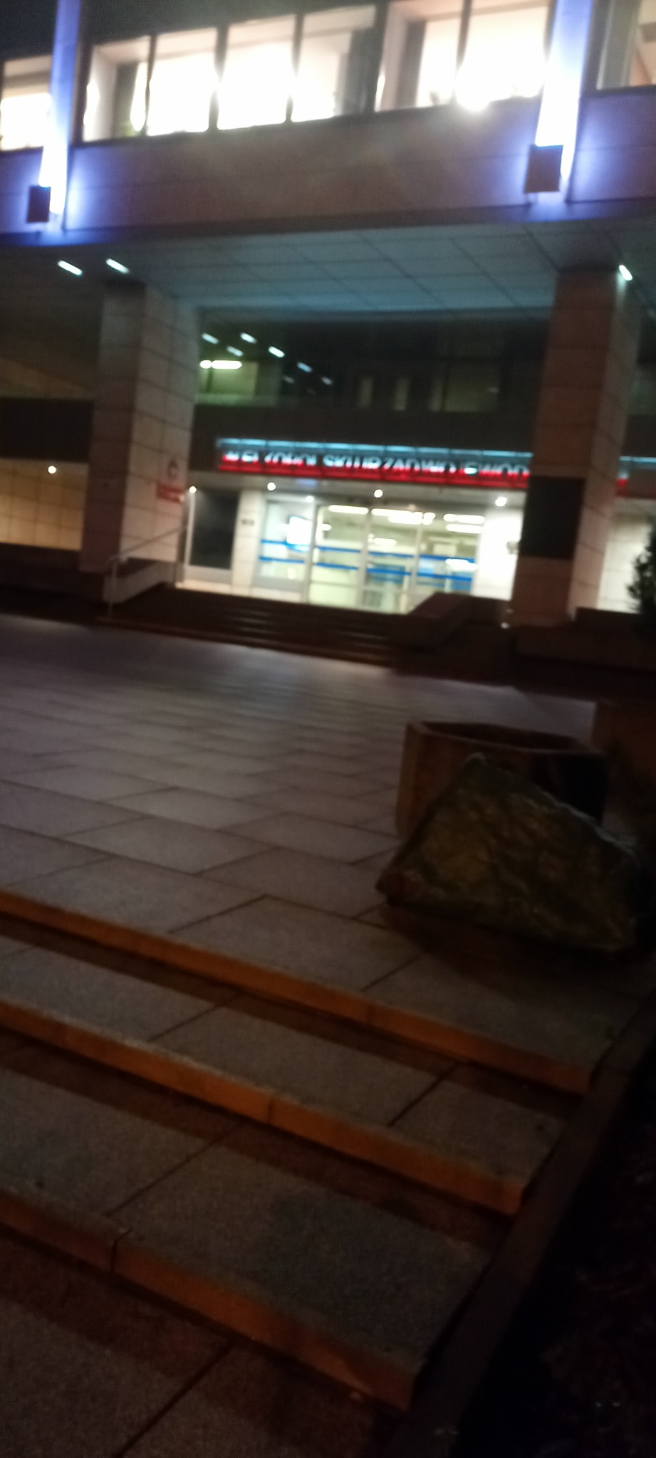 Nocne ujęcie z dołu na budynek z oświetlonymi oknami i kostką brukową. Widoczne schody i donica. Niska jakość zdjęcia.