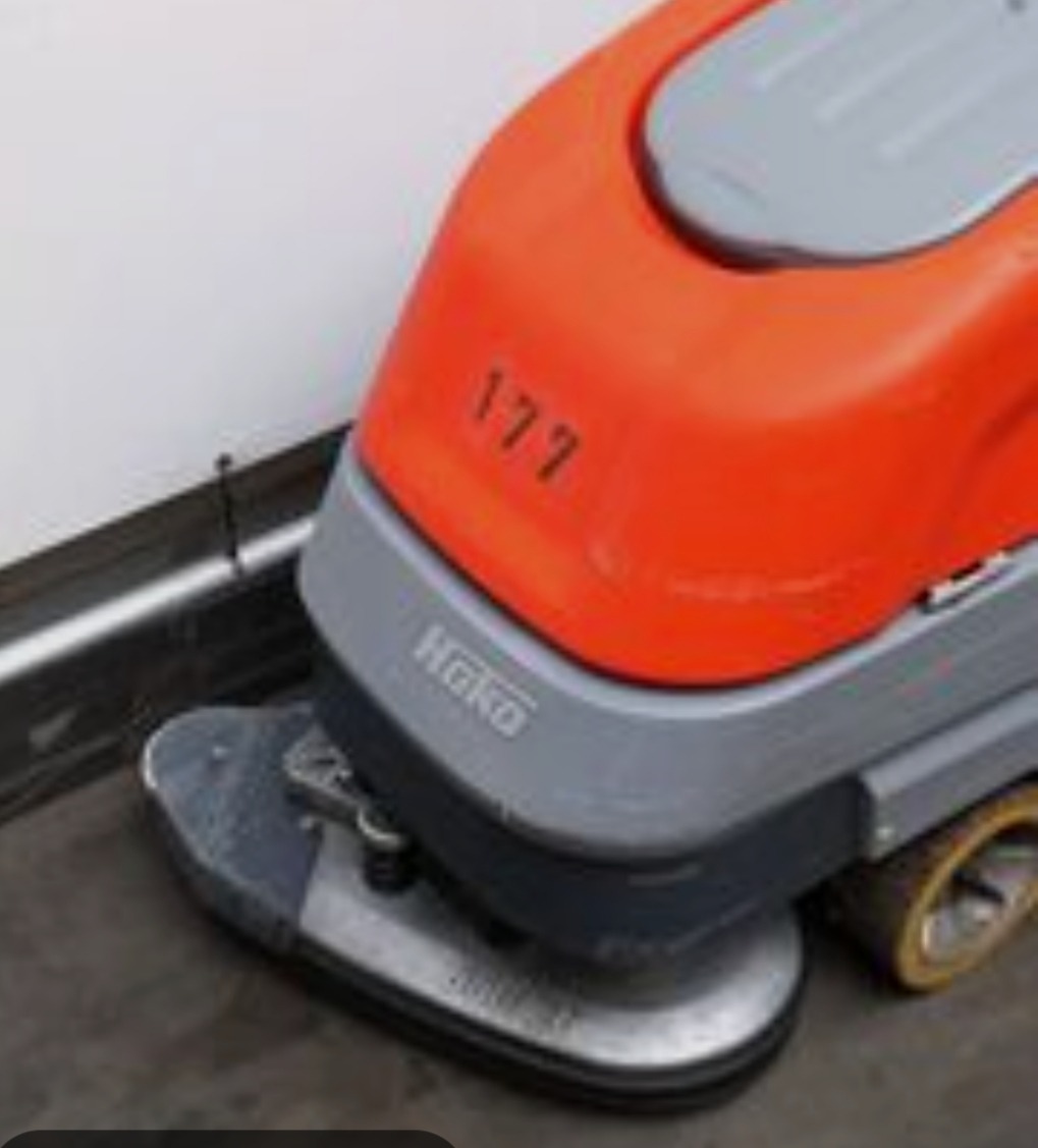 Automat szorująco-zbierający Hako w trakcie pracy na szarej posadzce, widoczne ślady wilgoci. Pomarańczowo-szara obudowa, szorstka powierzchnia czyszcząca.