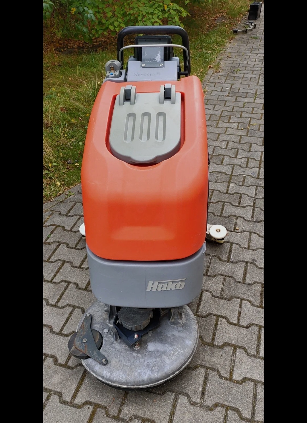 Pomarańczowo-szara maszyna Hako do czyszczenia kostki brukowej stoi na szarym bruku. Widok z góry na urządzenie z uchwytem i zbiornikiem.