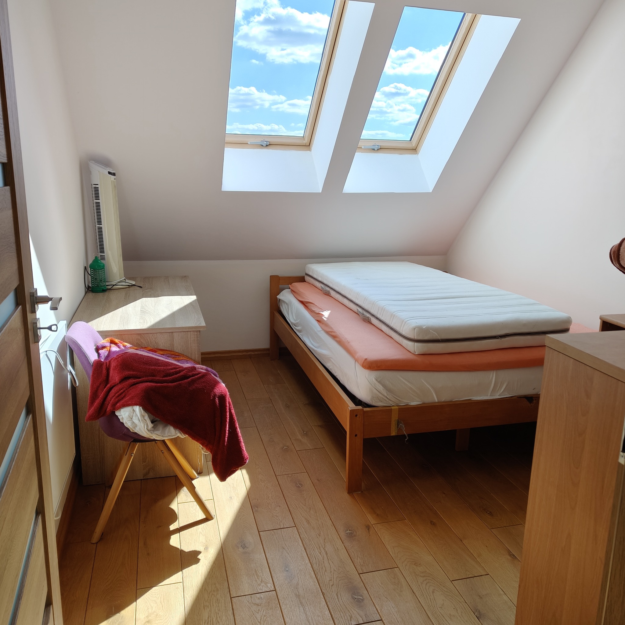 Pokój na poddaszu z drewnianą podłogą i oknami dachowymi. Widoczne łóżko, krzesło z kocem, grzejnik i szafka. Słoneczny dzień.