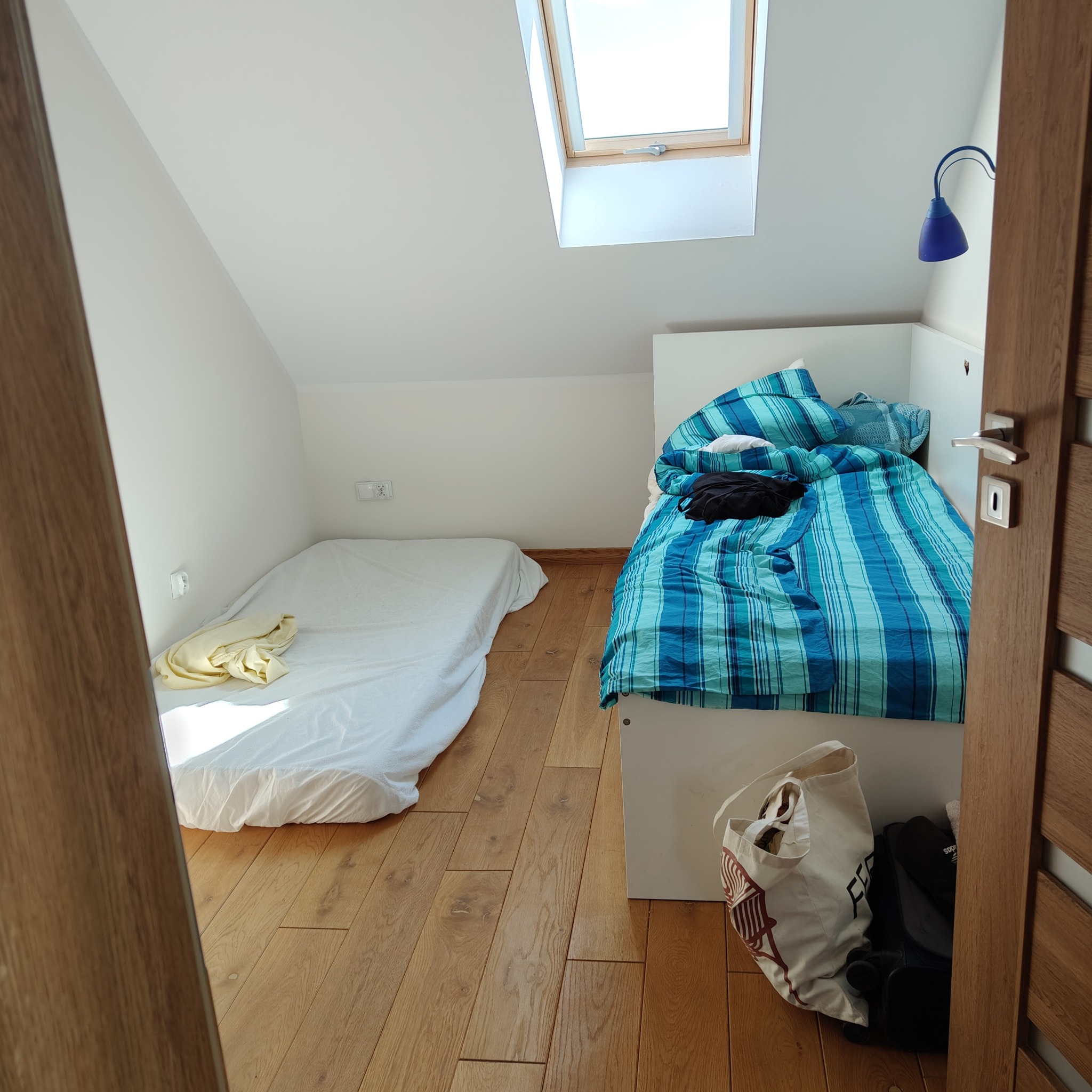 Pokój na poddaszu z dwoma łóżkami: jedno z niebieską pościelą w paski, drugie z białą. Widoczne okno dachowe i drewniana podłoga. Częściowo otwarte drzwi po prawej.
