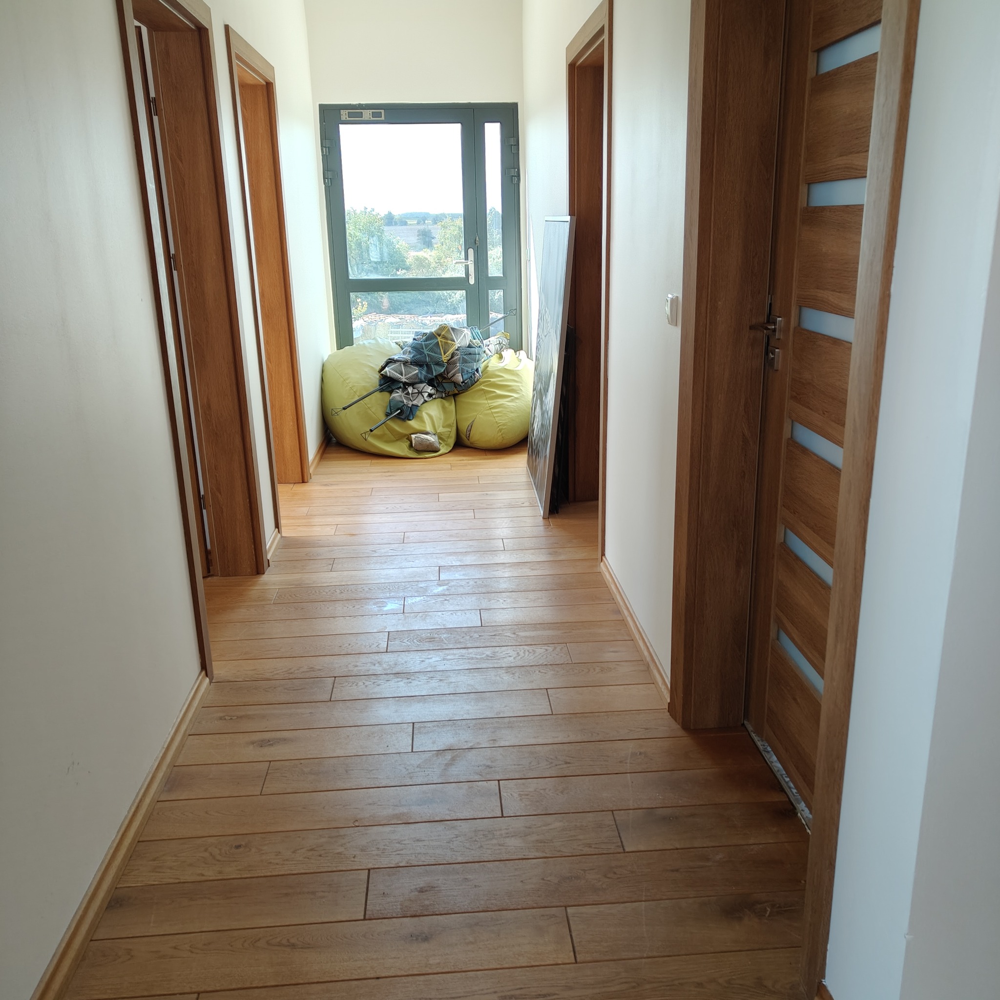 Korytarz z drewnianą podłogą i trzema parami drzwi. Dwa worki z resztkami po remoncie leżą przy oknie. Świeżo wykończone wnętrze po remoncie.