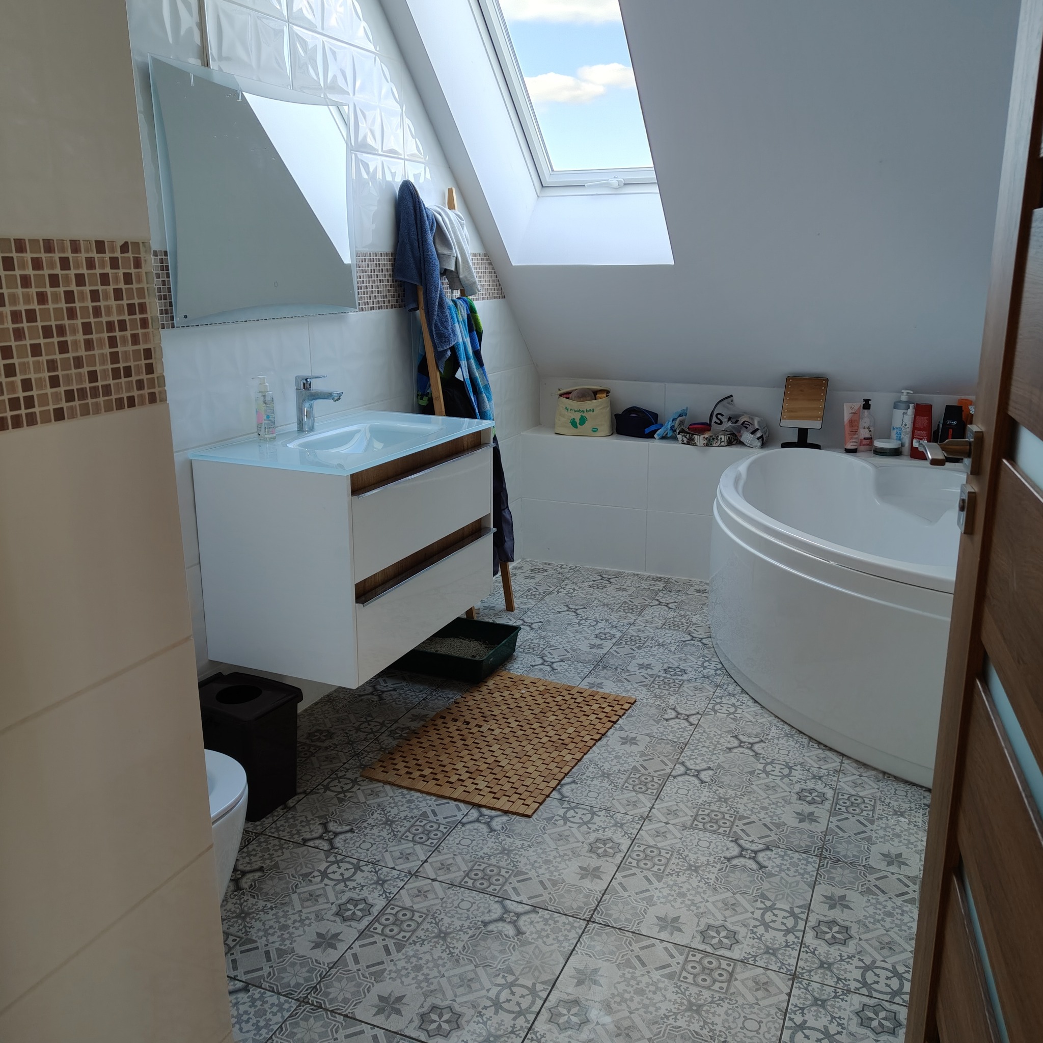 Jasna łazienka na poddaszu z oknem dachowym, białą wanną i umywalką z szafką. Na podłodze wzorzyste płytki, a na ścianach białe i mozaikowe płytki. Widoczne akcesoria łazienkowe.