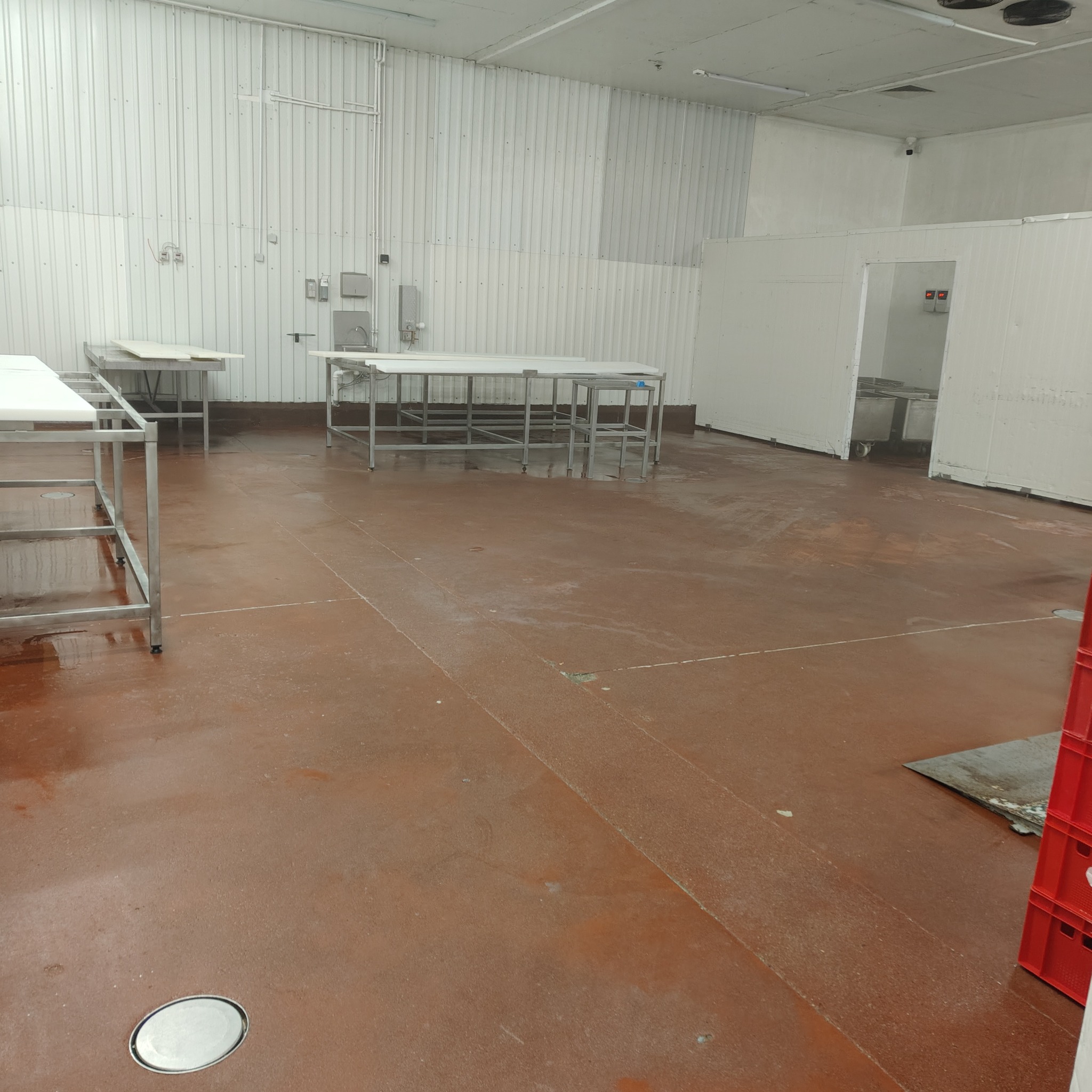 Wnętrze hali produkcyjnej z metalowymi stołami i brązową podłogą po myciu. Widoczne ślady wilgoci, białe ściany z blachy trapezowej i odpływ w podłodze.