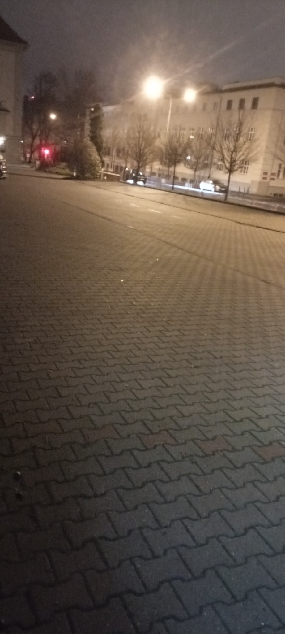 Nocne ujęcie z poziomu kostki brukowej, prowadzącej w kierunku oświetlonej ulicy z drzewami i budynkami w tle. Widoczne światła samochodów i latarni.