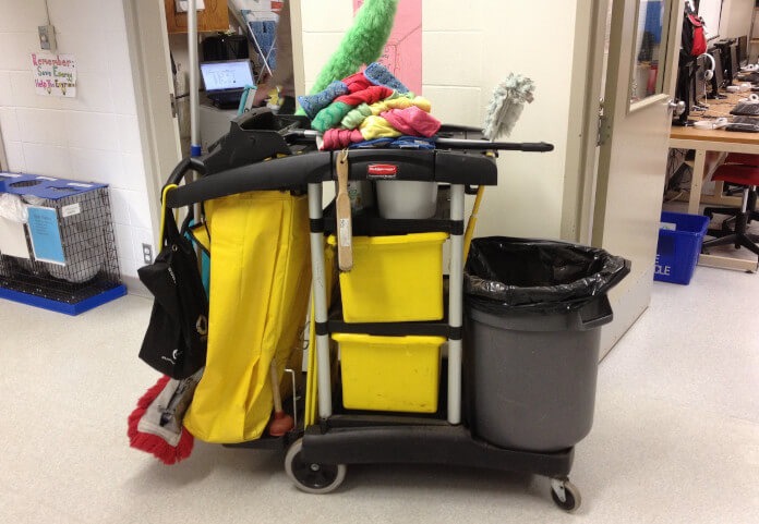 Wózek do sprzątania z mopem, ścierkami i pojemnikami na środki czystości w pomieszczeniu z biurkami i komputerami. Widoczny kosz na śmieci i żółte pojemniki.