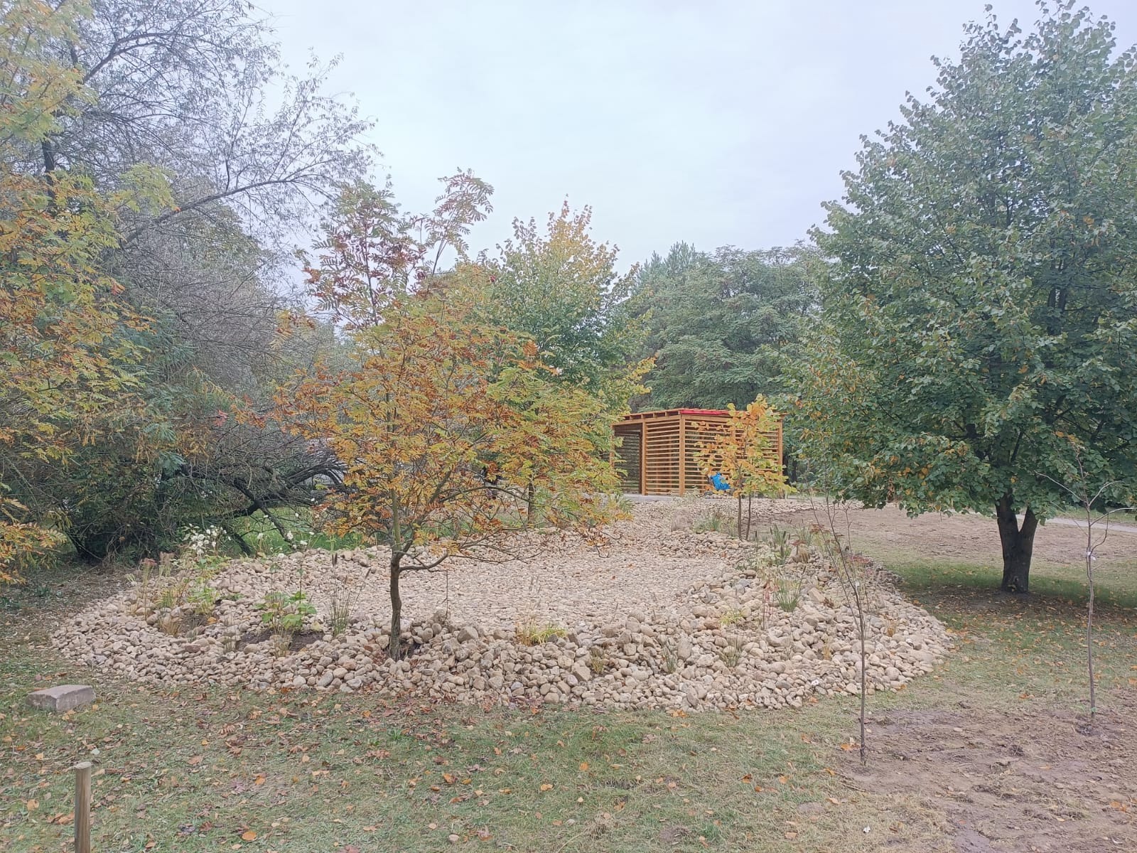 Jesienny krajobraz z drewnianą altaną na wzgórzu usypanym z kamieni, otoczonym drzewami z liśćmi w odcieniach zieleni, żółci i brązu. Pochmurne niebo w tle.