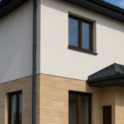 Elewacja dwukondygnacyjnego domu z beżową okładziną imitującą kamień, ciemnymi oknami i rynnami. Widok na narożnik budynku i fragment dachu.