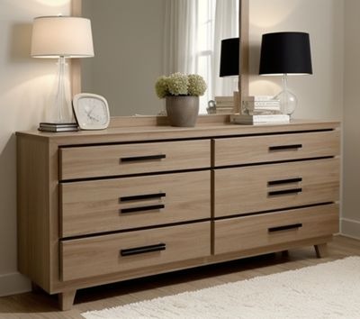 Drewniana komoda z sześcioma szufladami i czarnymi uchwytami, zwieńczona lustrem, lampami i dekoracjami. Naturalne oświetlenie podkreśla fakturę drewna.