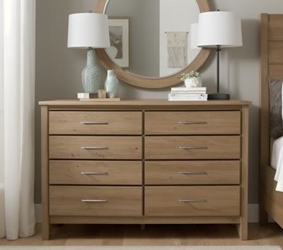 Jasna, drewniana komoda z ośmioma szufladami i srebrnymi uchwytami. Nad komodą okrągłe lustro w drewnianej ramie, lampki i dekoracje. Widoczny fragment łóżka.