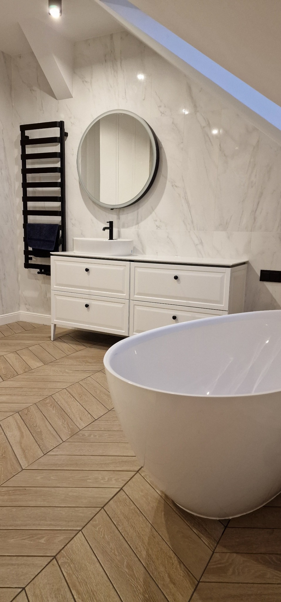 Nowoczesna łazienka na poddaszu: biała wanna wolnostojąca, szafka z umywalką, okrągłe lustro i marmurowe płytki ścienne. Parkiet ułożony w jodełkę.