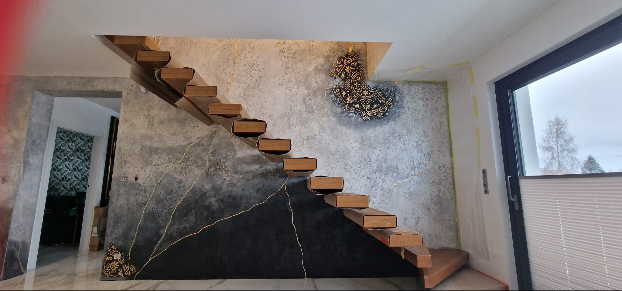 Nowoczesne schody z drewnianymi stopniami na tle ściany z efektem betonu i złotymi żyłkami, zdobione czarnym trójkątem i ornamentem. Widok na okno i wnętrze domu.