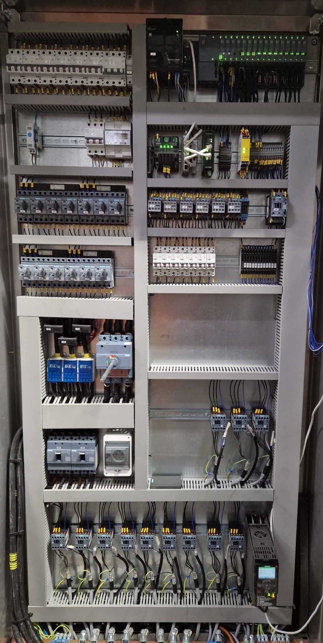 Szara szafa sterownicza z widocznymi bezpiecznikami, przekaźnikami i okablowaniem. Układ modułowy z elementami elektrycznymi i automatyki. Widoczne oznaczenia i podłączenia.