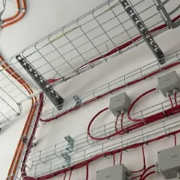 Instalacja kablowa: koryta siatkowe z przewodami w kolorze czerwonym i pomarańczowym, puszki instalacyjne na białej ścianie. Widok z dołu, oświetlenie naturalne.