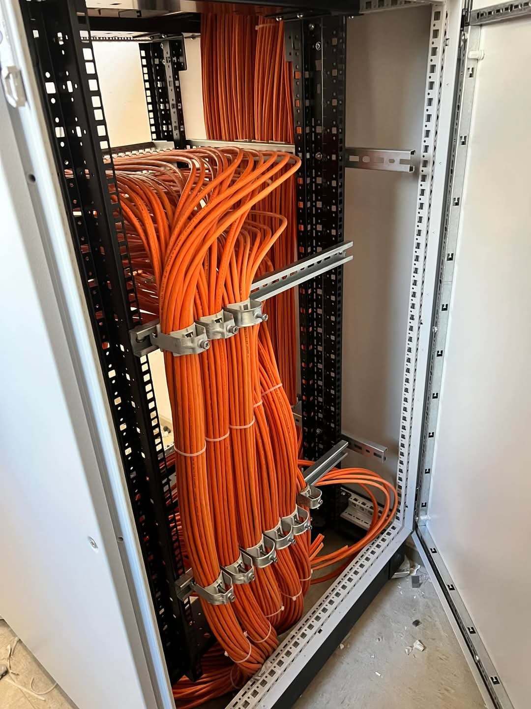 Pomarańczowe kable sieciowe uporządkowane w szafie rack, zabezpieczone metalowymi uchwytami. Widoczne elementy konstrukcyjne szafy i podłoga.