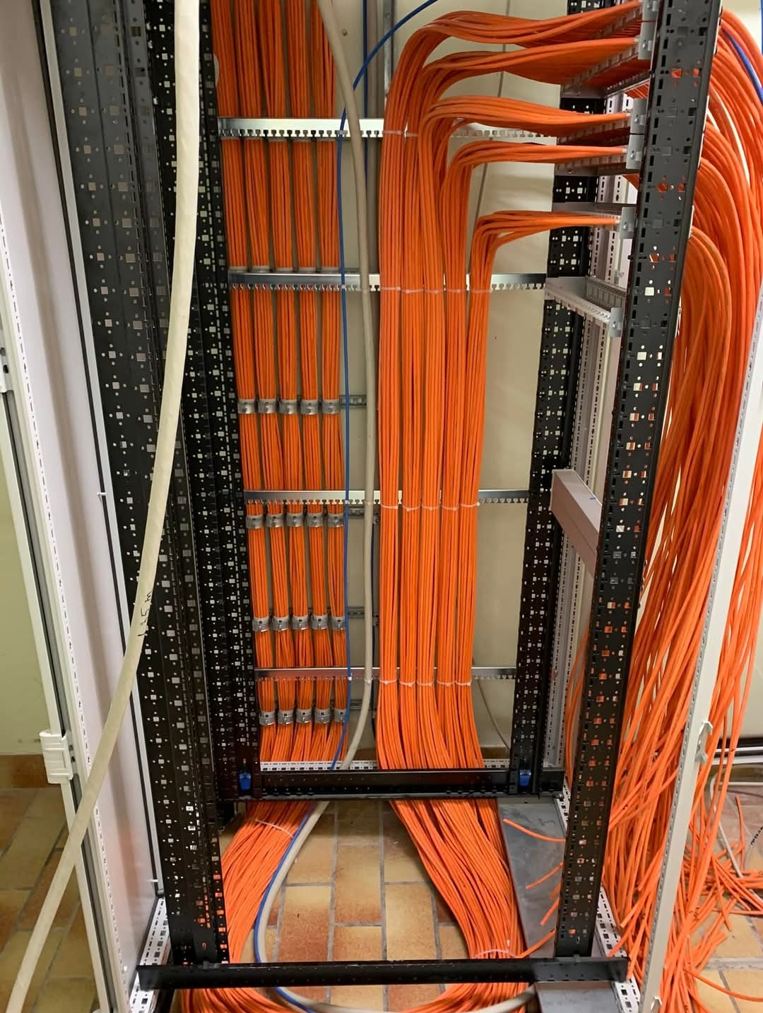 Pomarańczowe kable sieciowe uporządkowane w metalowej szafie rack, widoczne prowadnice kablowe i staranne ułożenie przewodów. Instalacja w serwerowni.