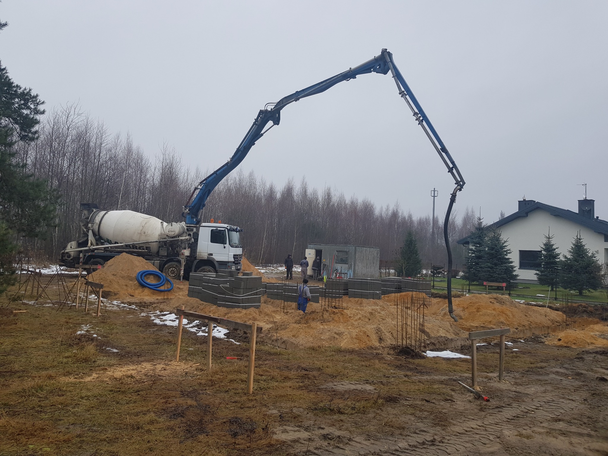 Betonomieszarka z pompą wylewa beton na fundamenty domu w trakcie budowy. Widoczni robotnicy w kombinezonach. W tle las i dom.