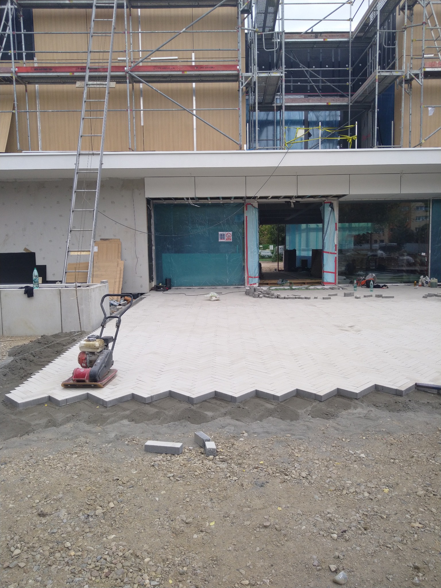 Układanie jasnej kostki w jodełkę na placu przed budynkiem w trakcie budowy. Widoczna ubijarka, częściowo ułożona kostka i przygotowane podłoże.