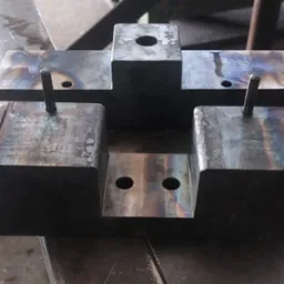 Metalowy element konstrukcyjny z otworami i pinami, przypominający formę odlewniczą. Widoczne ślady obróbki i przebarwienia termiczne na powierzchni.