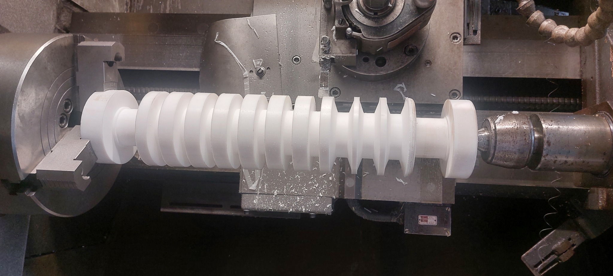 Biały, obrabiany element z tworzywa sztucznego na tokarce. Widoczne wióry i narzędzie tokarskie. Obróbka rowków.