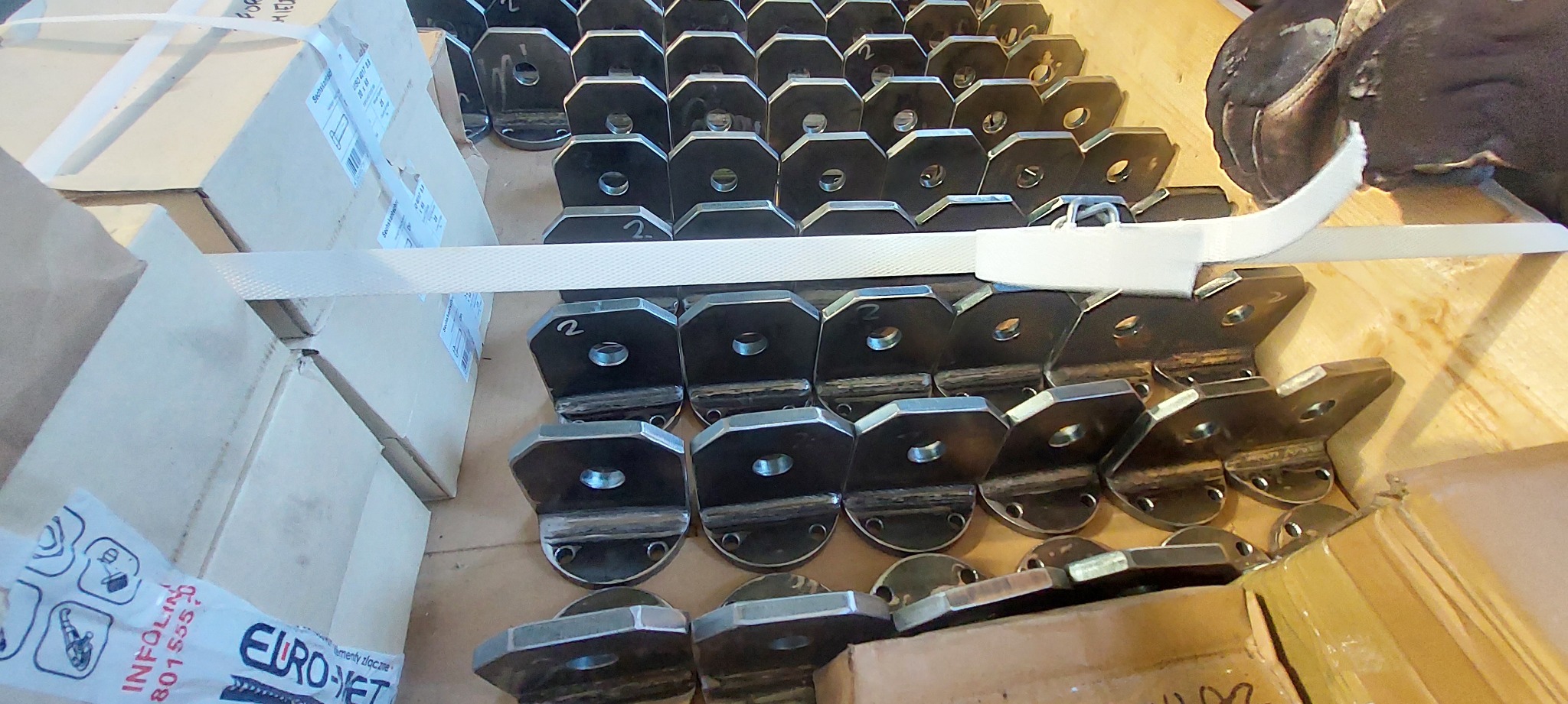 Szereg metalowych uchwytów, każdy z otworem i spawaną podstawą, ułożonych na palecie obok kartonów, zabezpieczonych taśmą. Na każdym elemencie widnieje numer '2'.