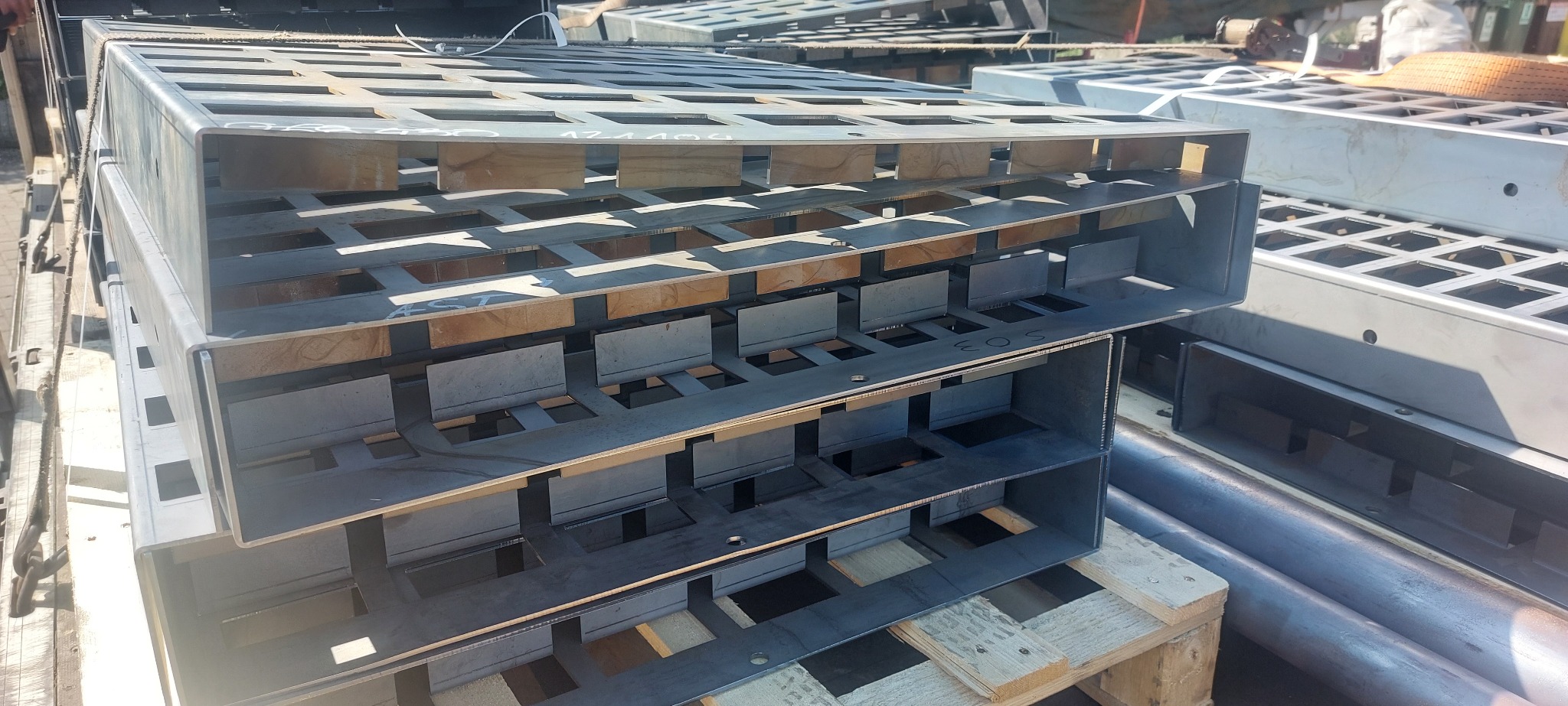 Stalowe konstrukcje z wyciętymi otworami, ułożone na palecie, widoczne numery seryjne. Elementy konstrukcyjne, wykonane z metalu, w surowym stanie.