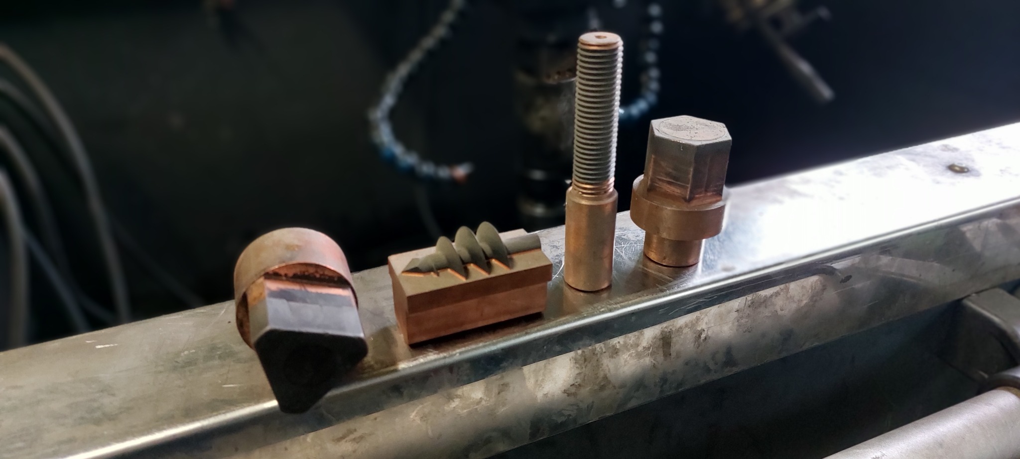 Miedziane elementy po obróbce skrawaniem: śruba, nakrętka i blok z wyfrezowanymi rowkami, ułożone na metalowej powierzchni w warsztacie.