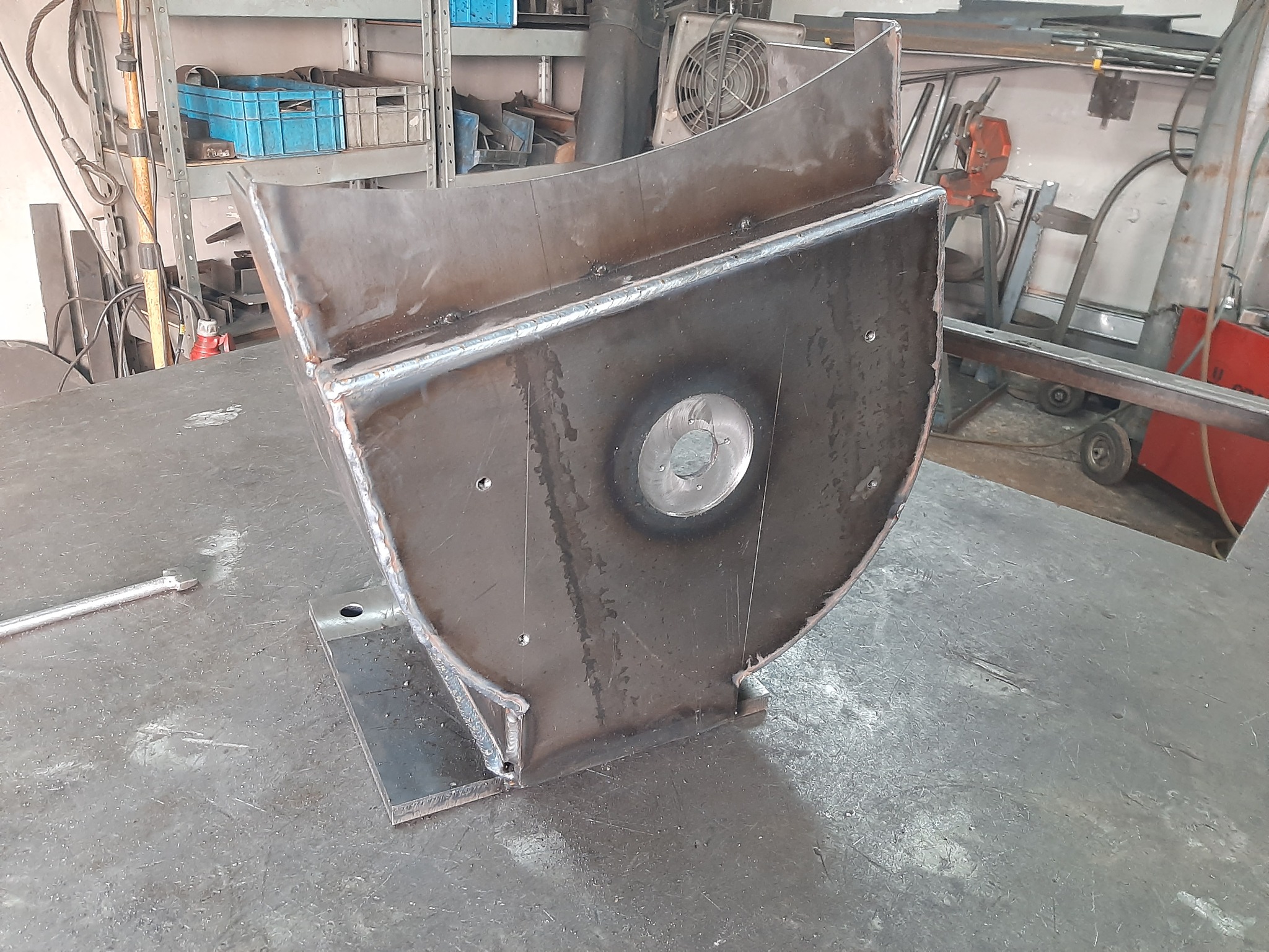 Spawana konstrukcja metalowa z otworem pośrodku, stojąca na stole warsztatowym. Widoczne ślady spawania i obróbki metalu. W tle warsztat z narzędziami.