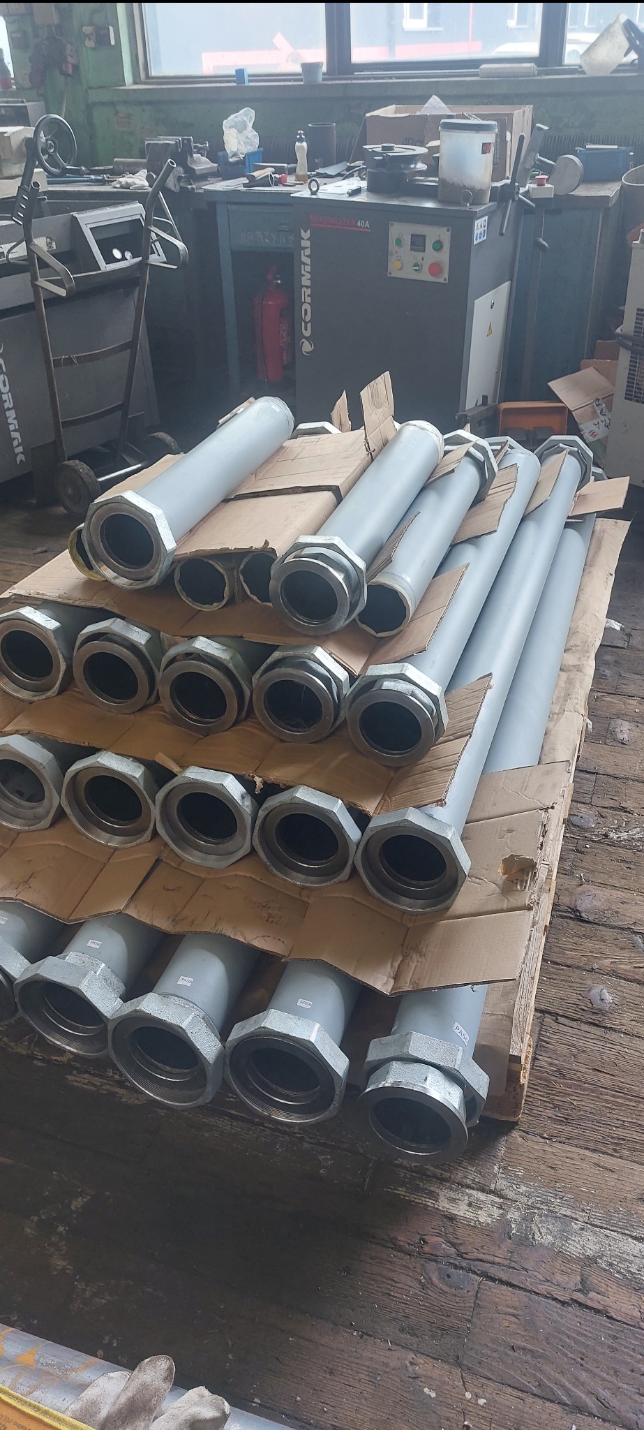 Szare, metalowe rury z łącznikami ułożone na palecie w warsztacie. Widoczne maszyny i narzędzia w tle. Rury przygotowane do montażu.
