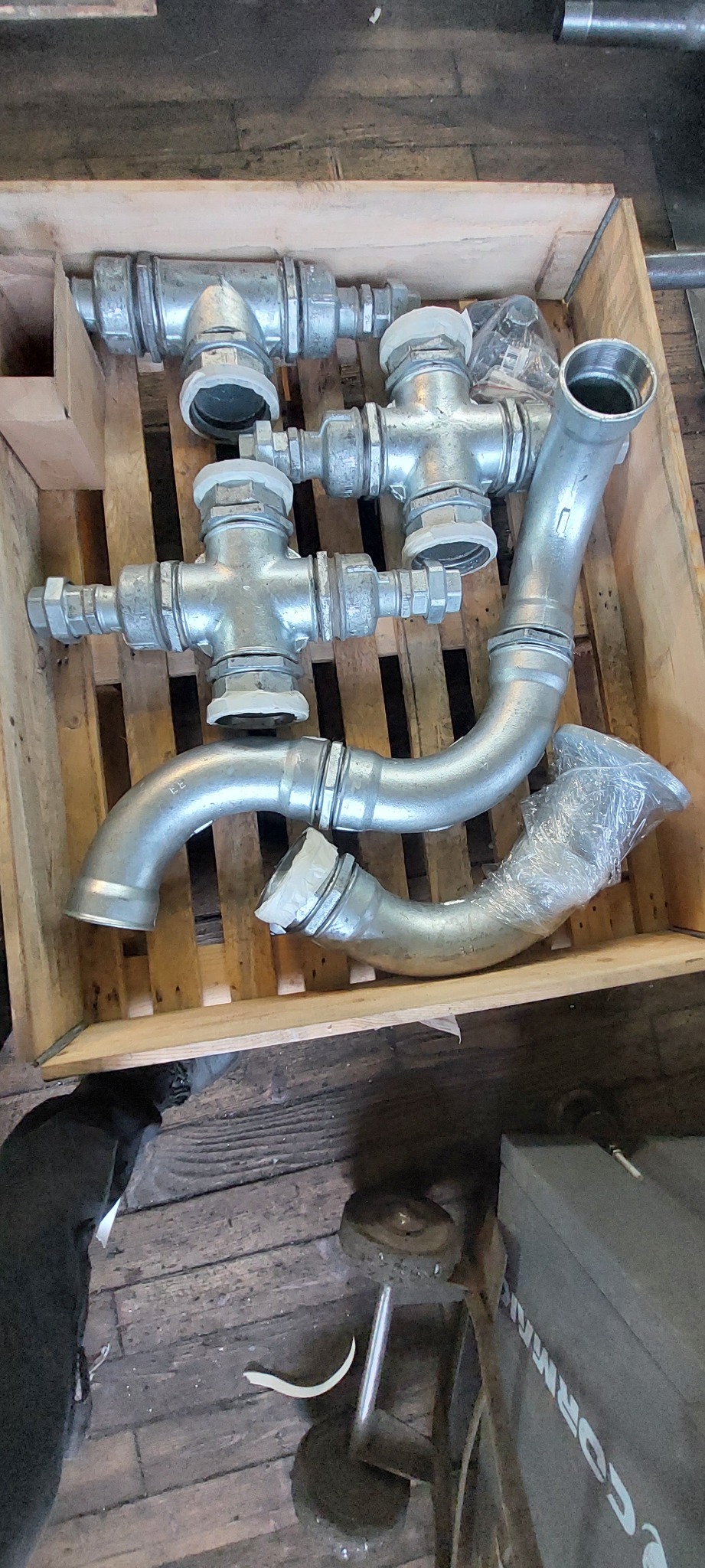 Metalowe kształtki rurowe (kolana, trójniki) w drewnianej skrzyni transportowej, częściowo zabezpieczone folią. Widoczne ślady użytkowania na elementach.