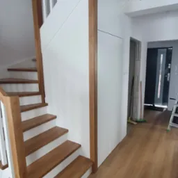 Biało-drewniane schody z balustradą, widok z dołu, jasne wnętrze, drewniana podłoga, minimalistyczny design, częściowo widoczne drzwi w tle.