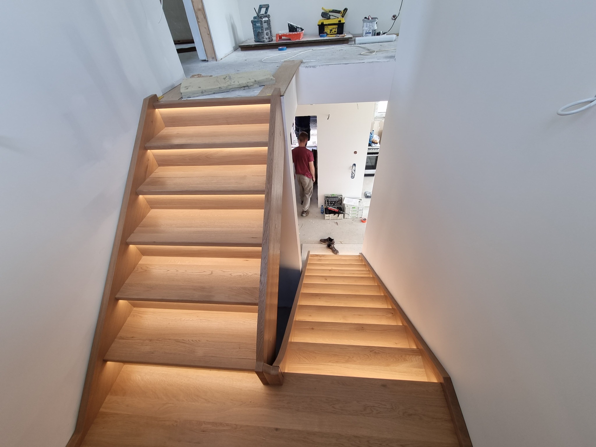 Nowoczesne schody drewniane z subtelnym podświetleniem LED, prowadzące na wyższą kondygnację. Widoczny fragment remontowanego pomieszczenia w tle.