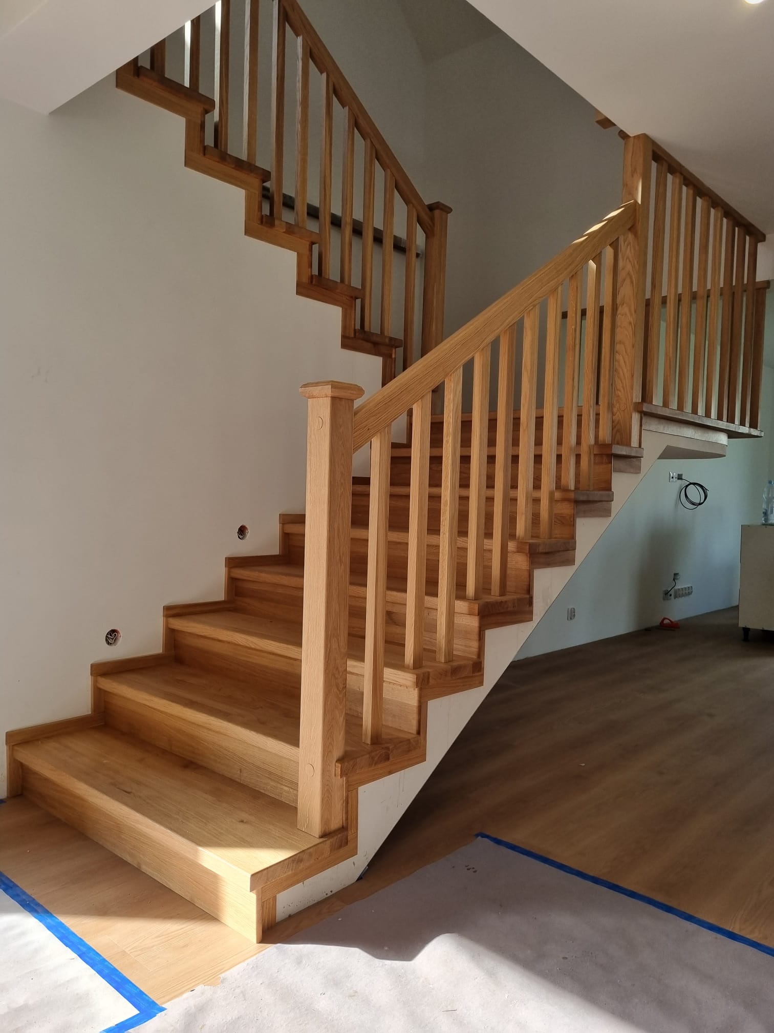 Jasne, drewniane schody z balustradą w nowoczesnym wnętrzu. Widoczne zabezpieczenia podłogi, podkreślające etap prac wykończeniowych.