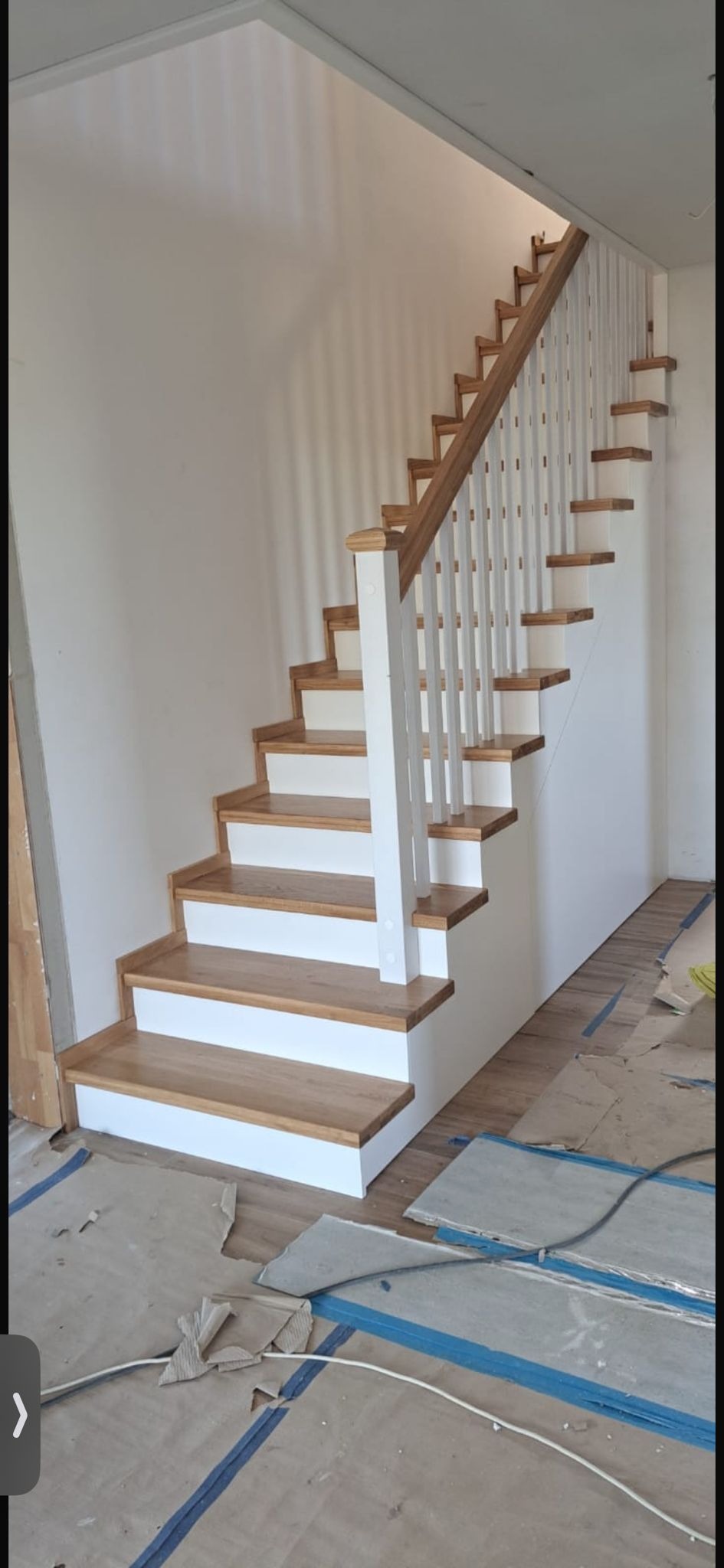 Klasyczne schody z jasnego drewna z białymi podstopnicami i balustradą, widok z dołu, w tle białe ściany. Częściowo zabezpieczona podłoga.