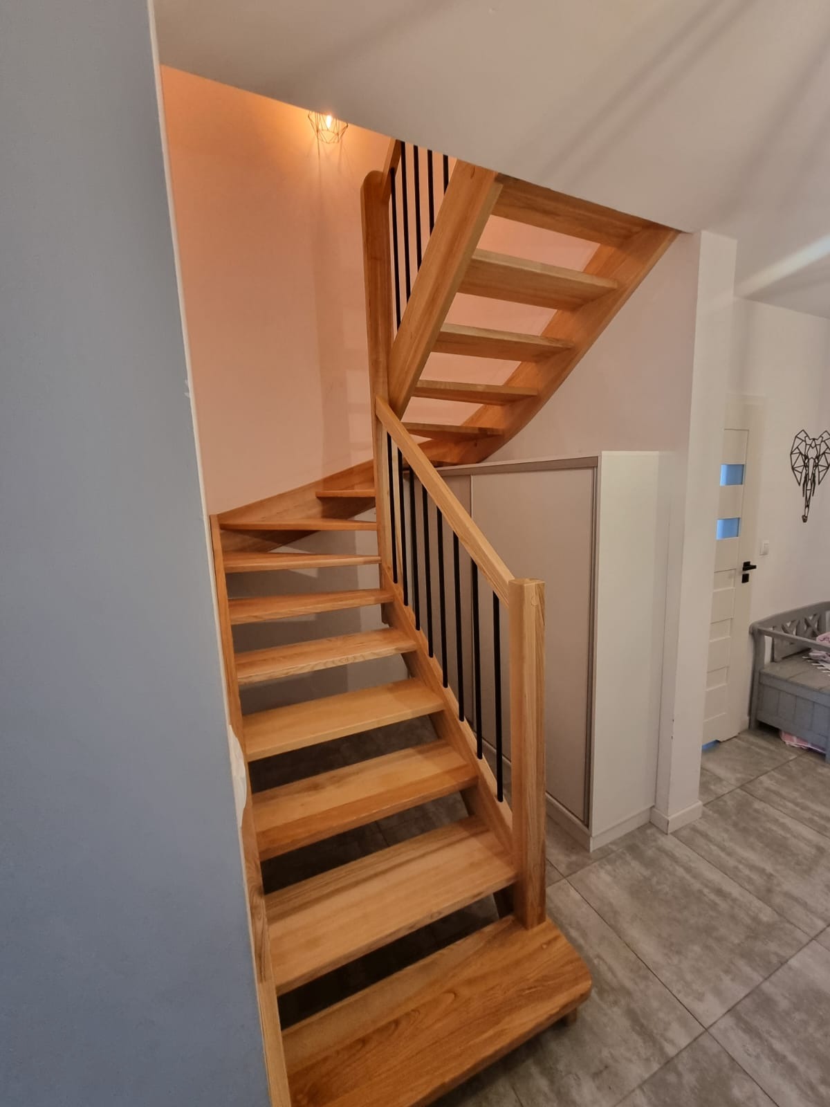 Jasne, drewniane schody z czarnymi balustradami w nowoczesnym wnętrzu. Widok z dołu, ukazujący zakręt schodów i detale wykończenia.