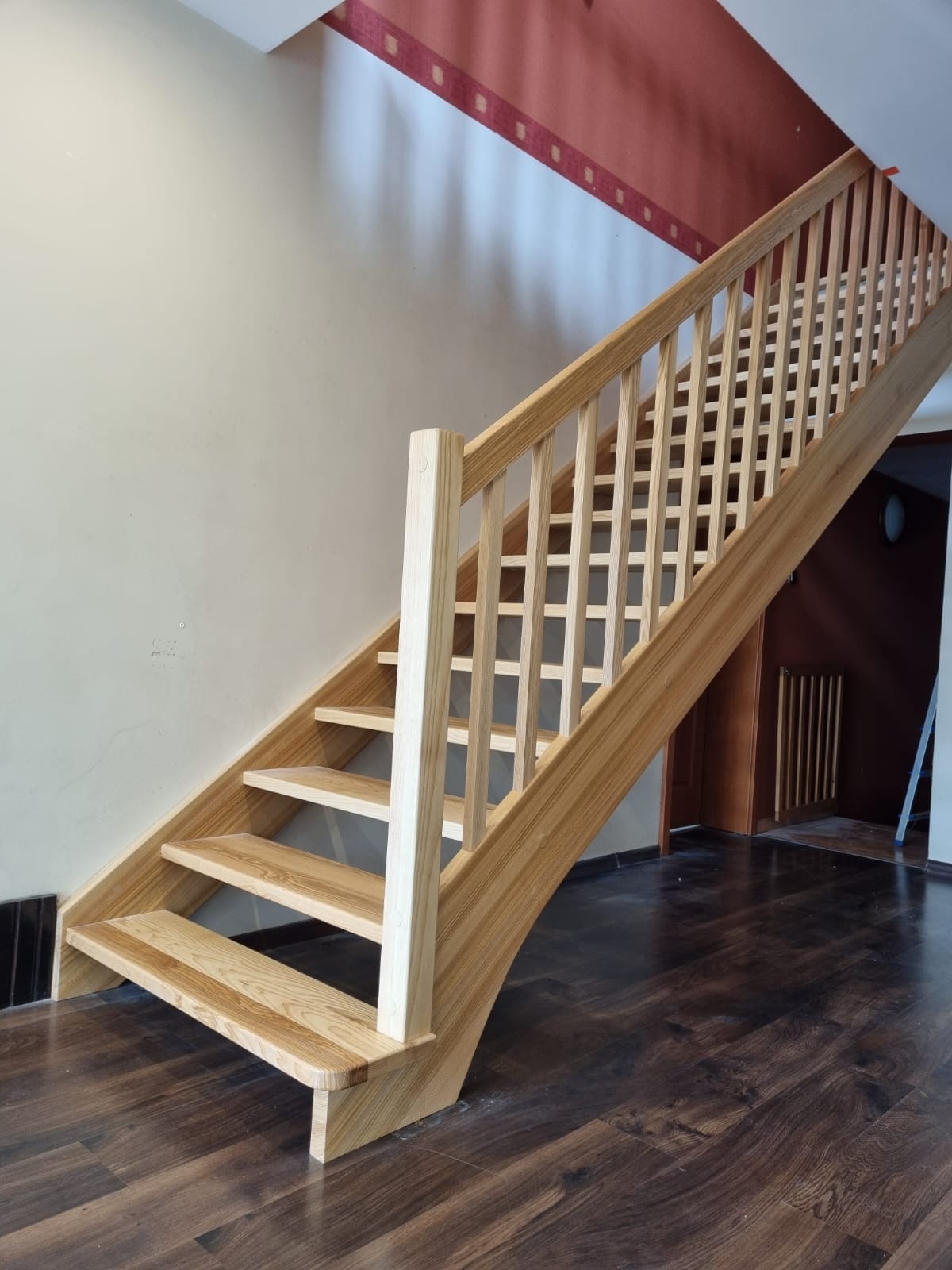 Jasne, ażurowe schody z drewna o naturalnym odcieniu, z widocznym rysunkiem słojów, prowadzące na piętro. Ciemna podłoga kontrastuje z kolorem schodów.