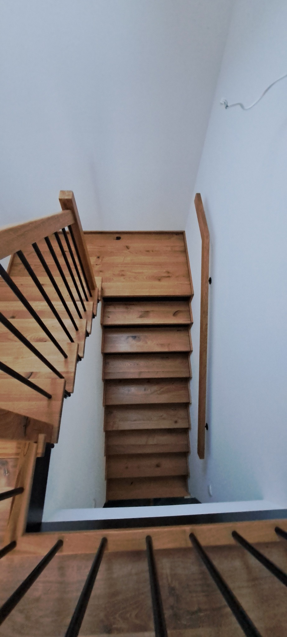 Naturalne, jasne schody z drewna, widok z góry, z czarnymi poręczami. Prosty bieg schodów przy białej ścianie. Minimalistyczny design.