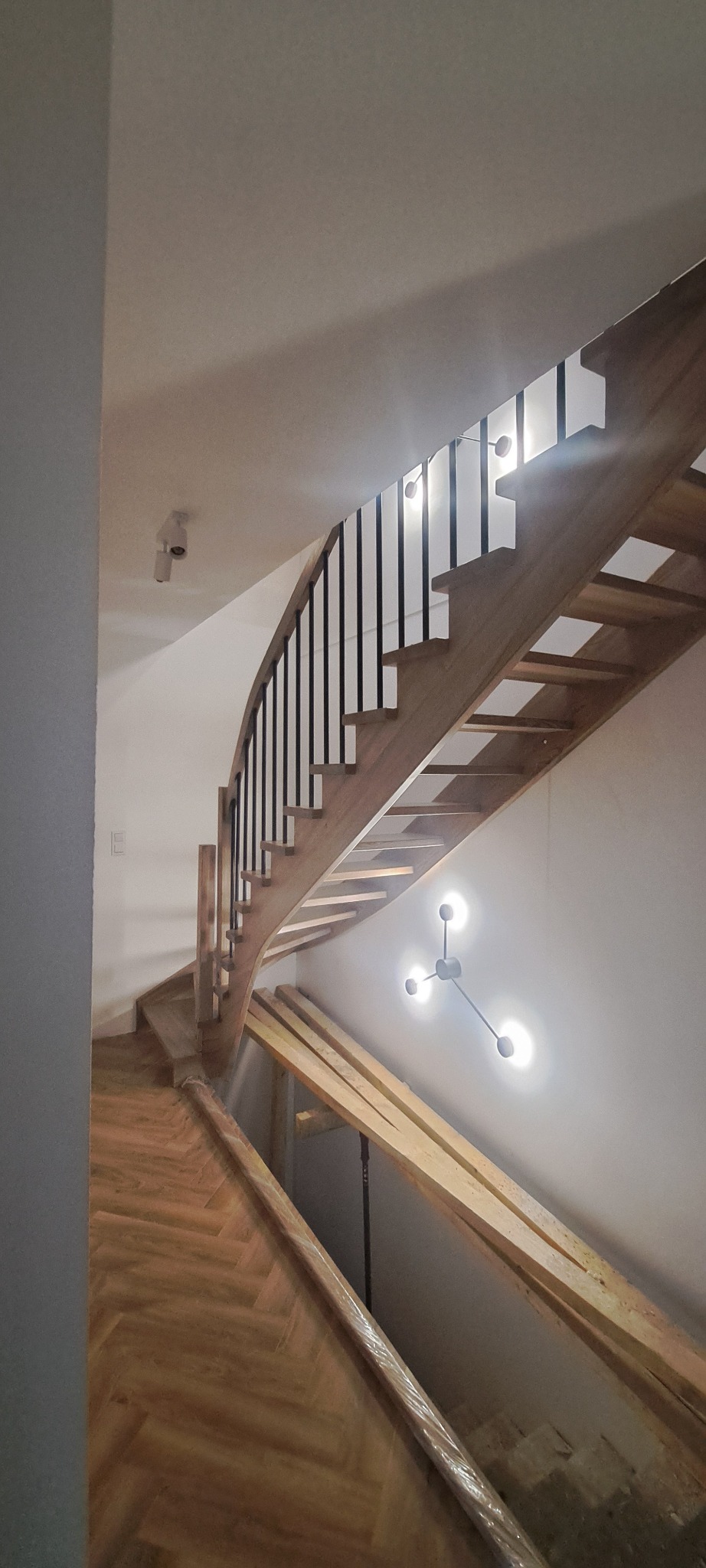 Zakrzywione schody z drewnianymi stopniami i czarną balustradą, oświetlone lampą w stylu molekularnym. Widoczne zabezpieczenia na podłodze.