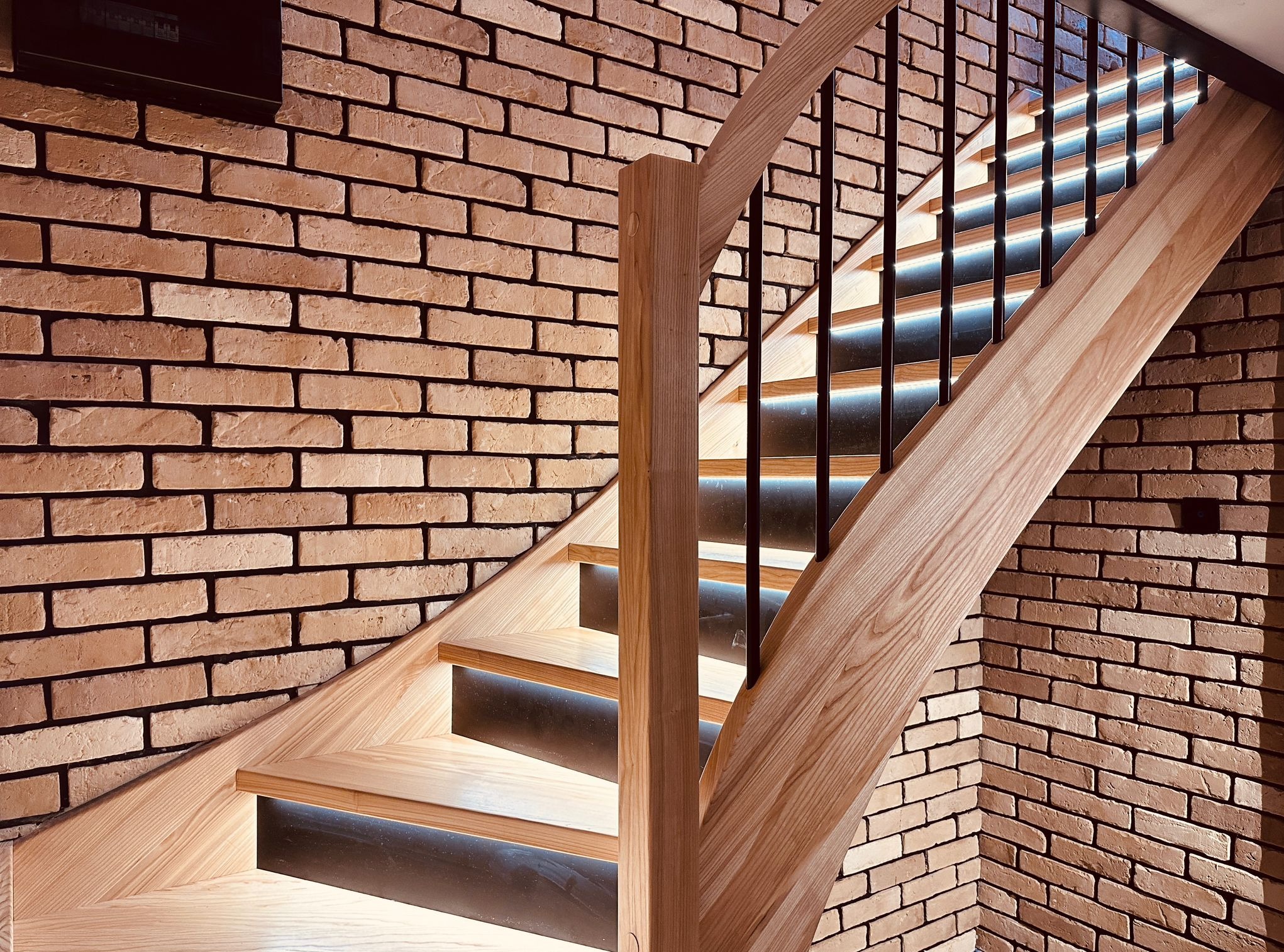 Nowoczesne schody drewniane z podświetleniem LED, czarne balustrady, ceglana ściana w tle. Widok z dołu, podkreślający detale wykończenia i oświetlenie.