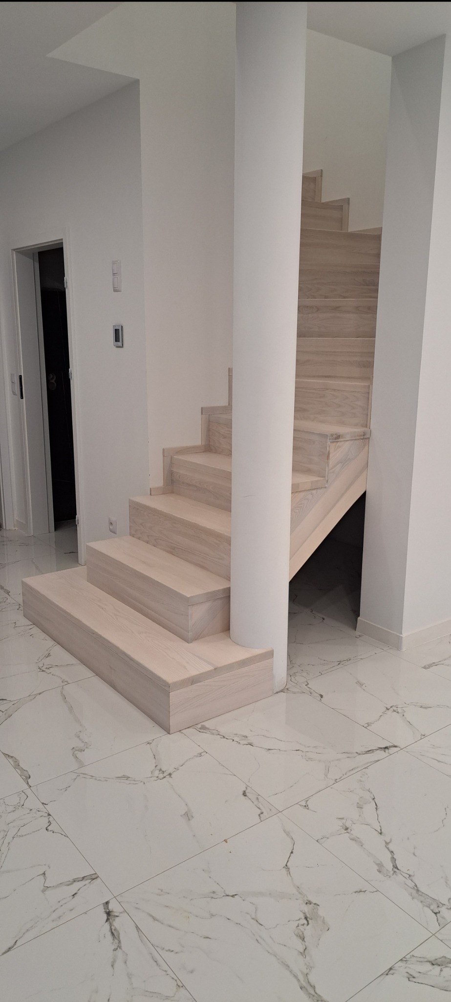 Jasne, minimalistyczne schody z drewna jesionowego owijające filar w nowoczesnym wnętrzu z marmurową podłogą i białymi ścianami.