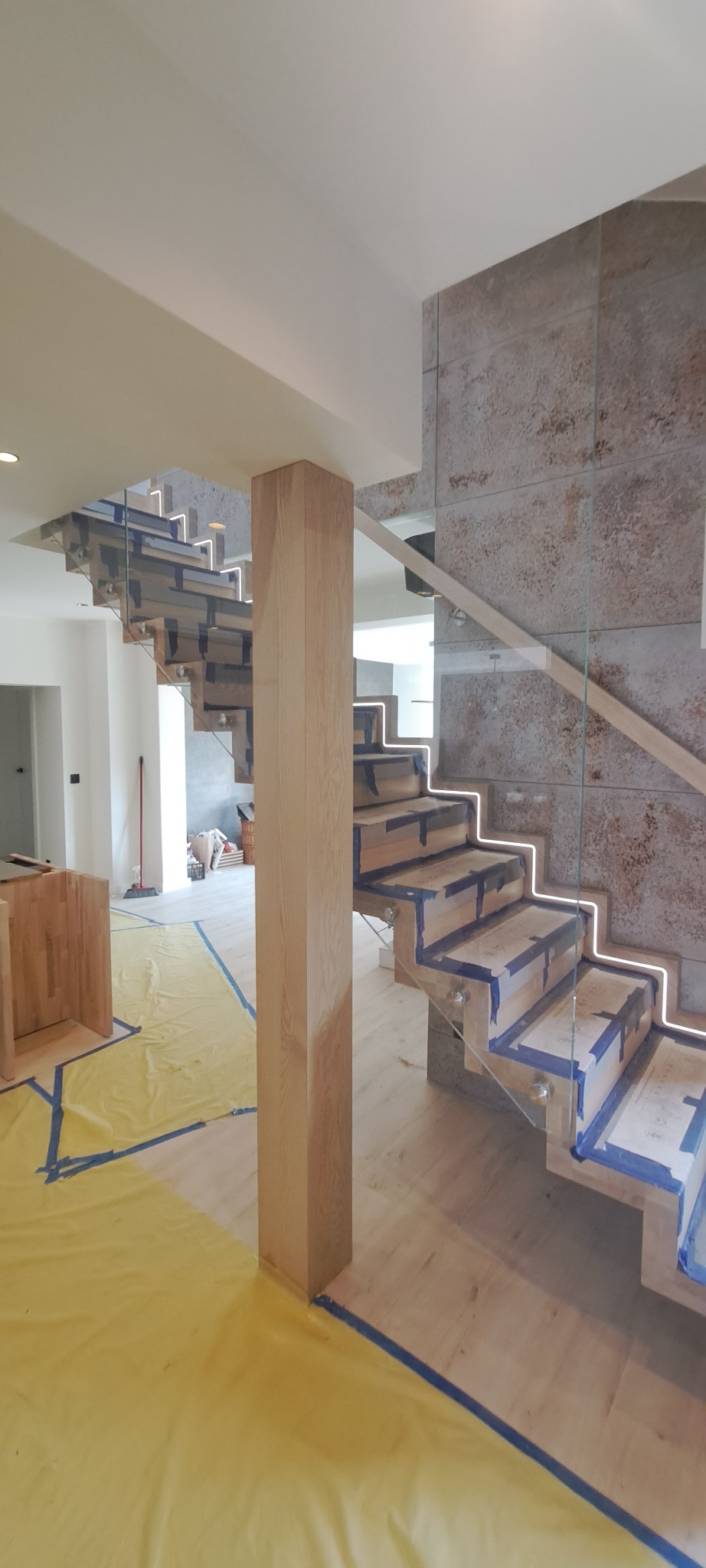 Nowoczesne schody z drewnianymi stopniami, szklaną balustradą i podświetleniem LED w trakcie montażu. Ochrona podłogi folią malarską.